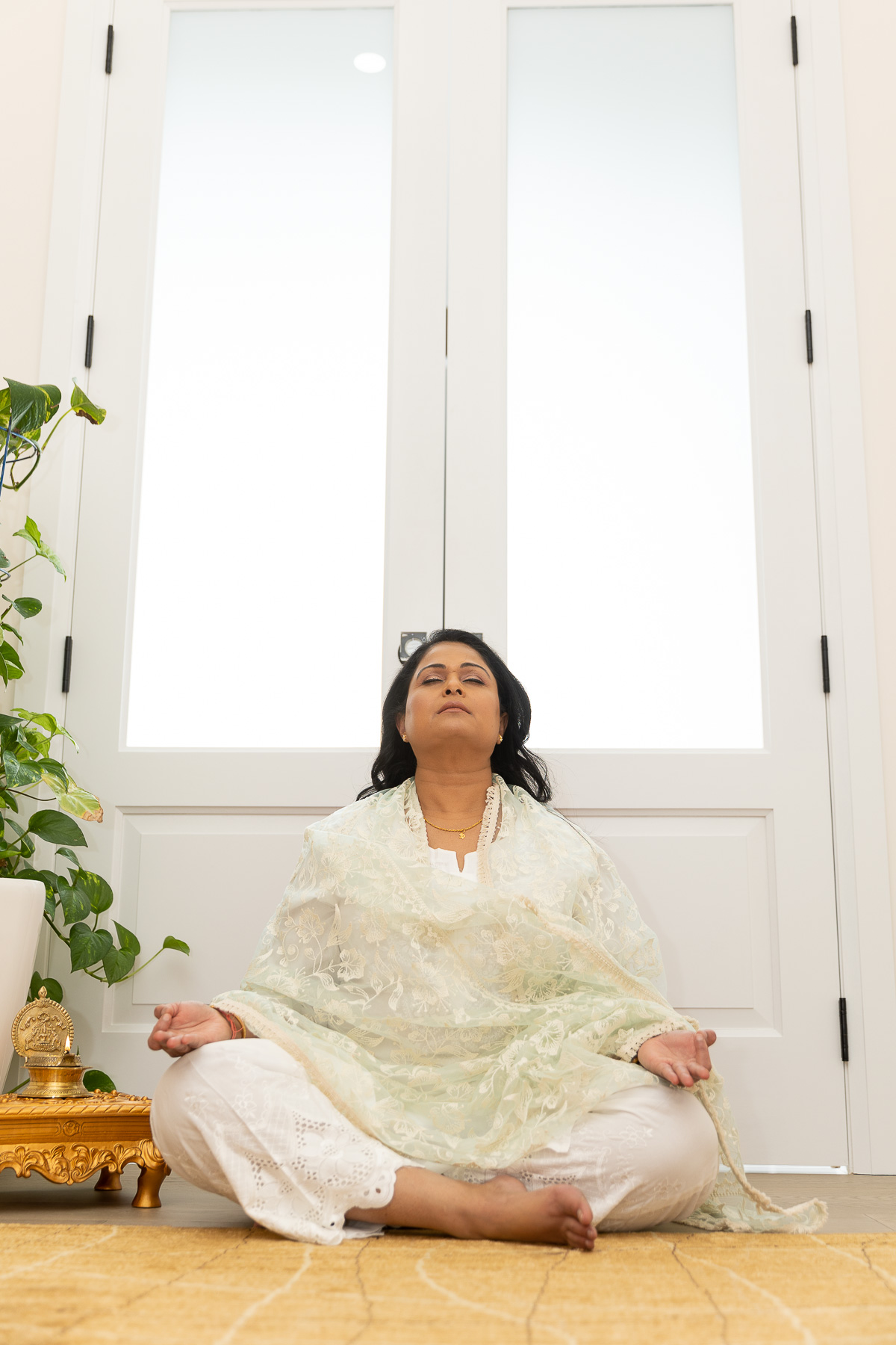 Kalinie meditating with her legs crossed, looking up towards the ceiling. She is in light coloured Indian clothing on a yellow mat. Behind her is a door with windows letting in bright light