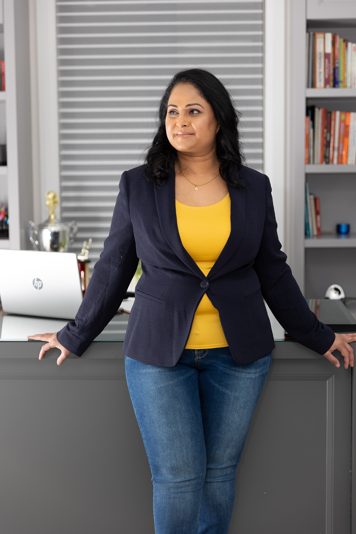 Kalinie, founder of Conscious Empowerment in her office wearing a blazer, jeans, and yellow shirt. She looks pensive and calm.